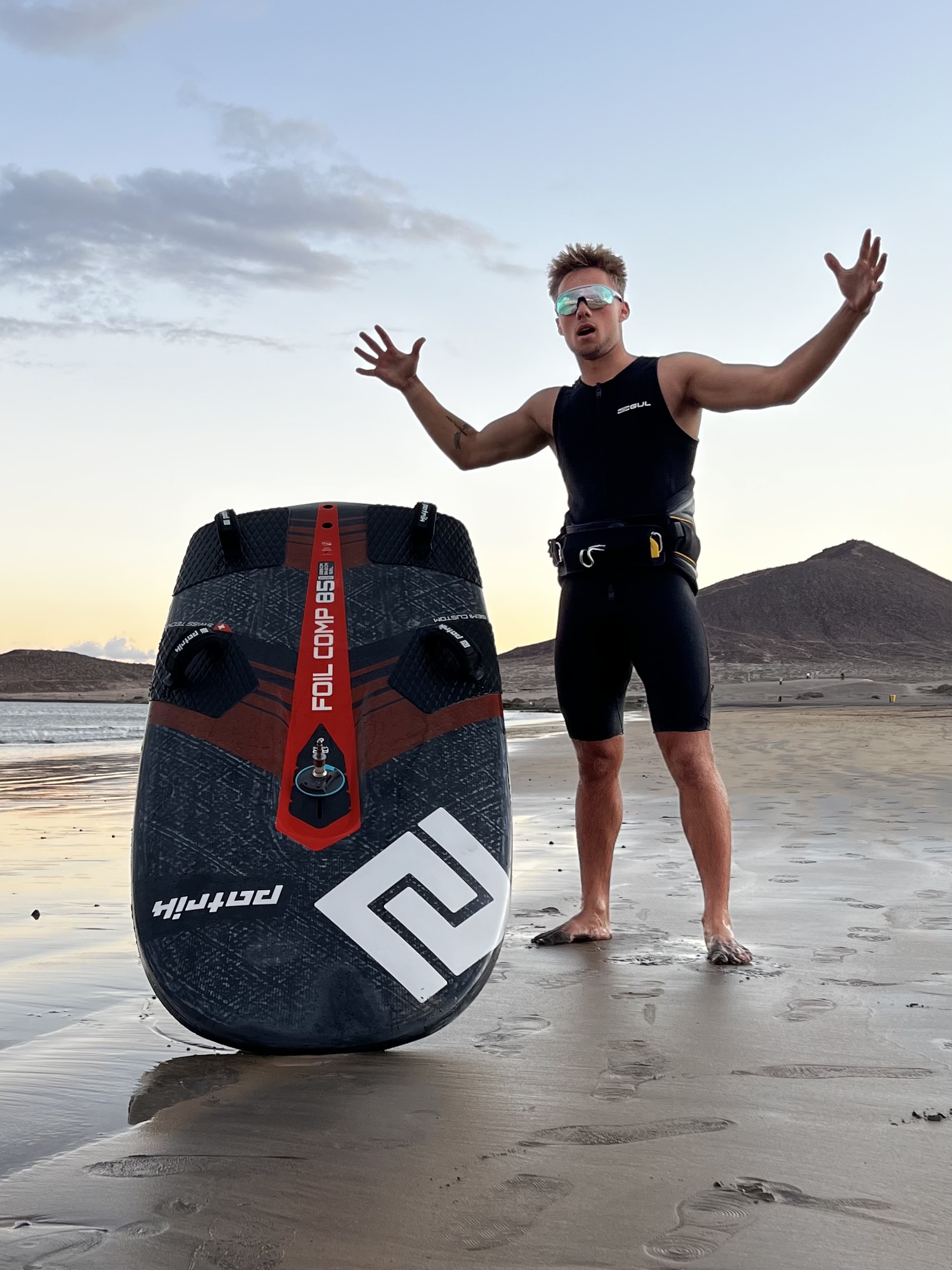 Noah with arms up on the beach next to his foil board
