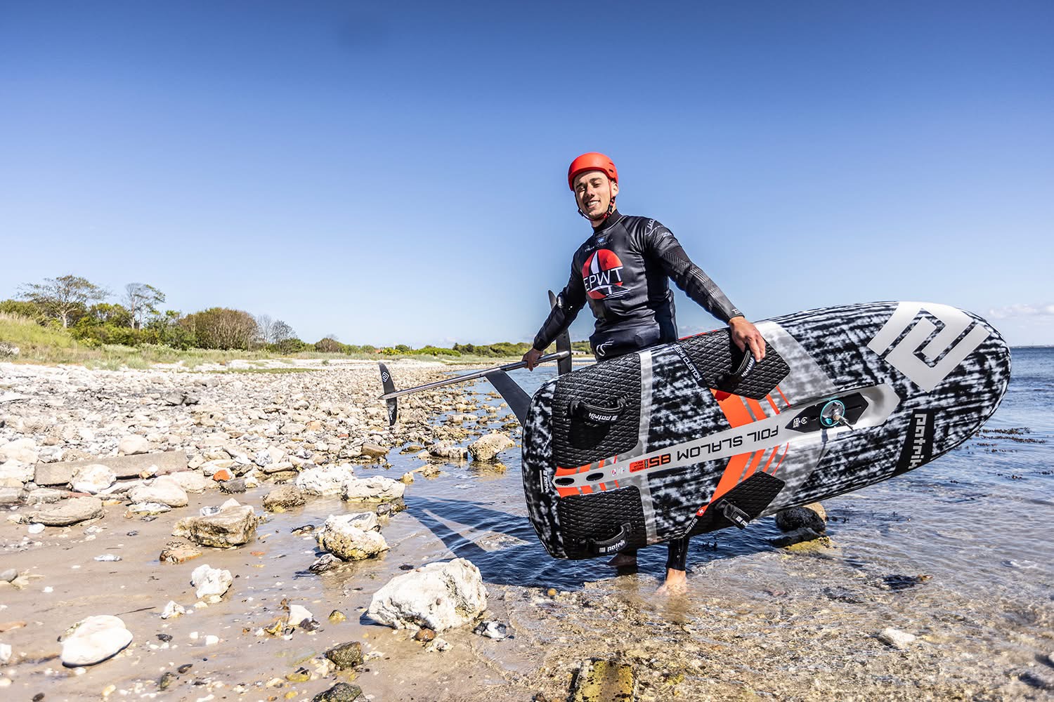 Noah in a red helmet wading ashore on a rocky beach, carrying his Patrik foil slalom board