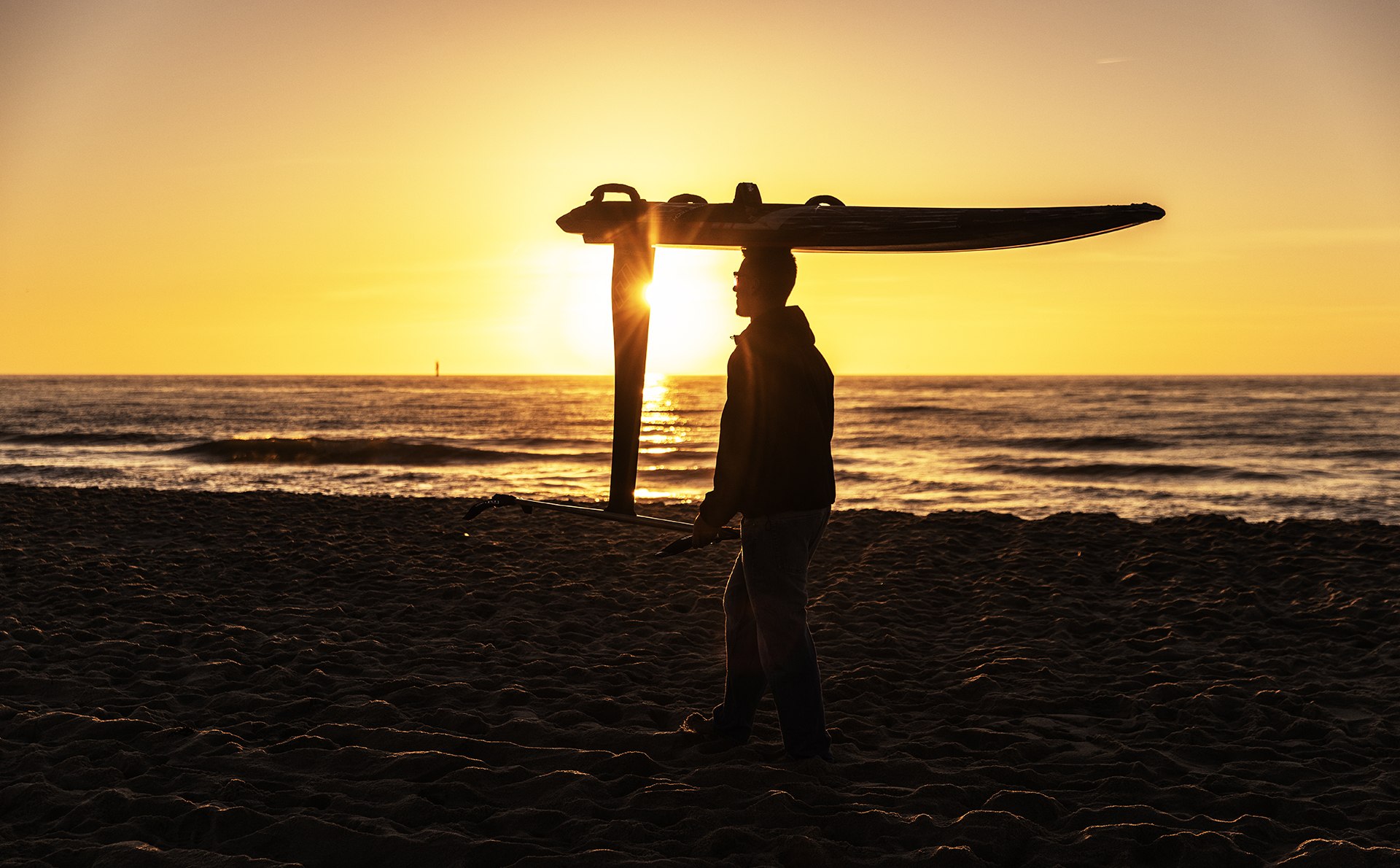 Silhouette of Noah carrying his foil board on his head on the beach at sunset