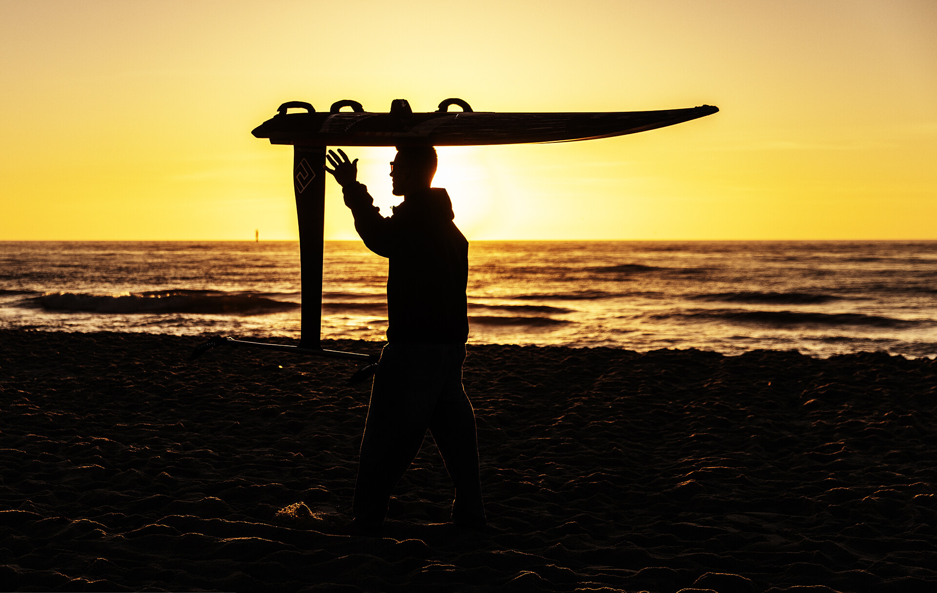 Silhouette of Noah at sunset on the beach, his foil board on his head and one hand raised