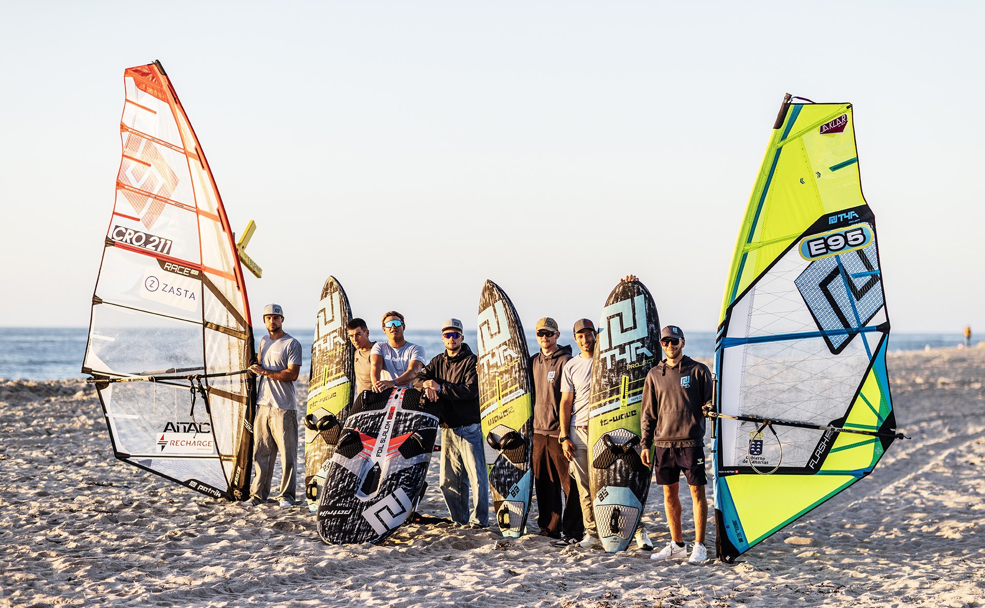 Noah with fellow riders on the beach at sunset, boards and sails standing in the sand